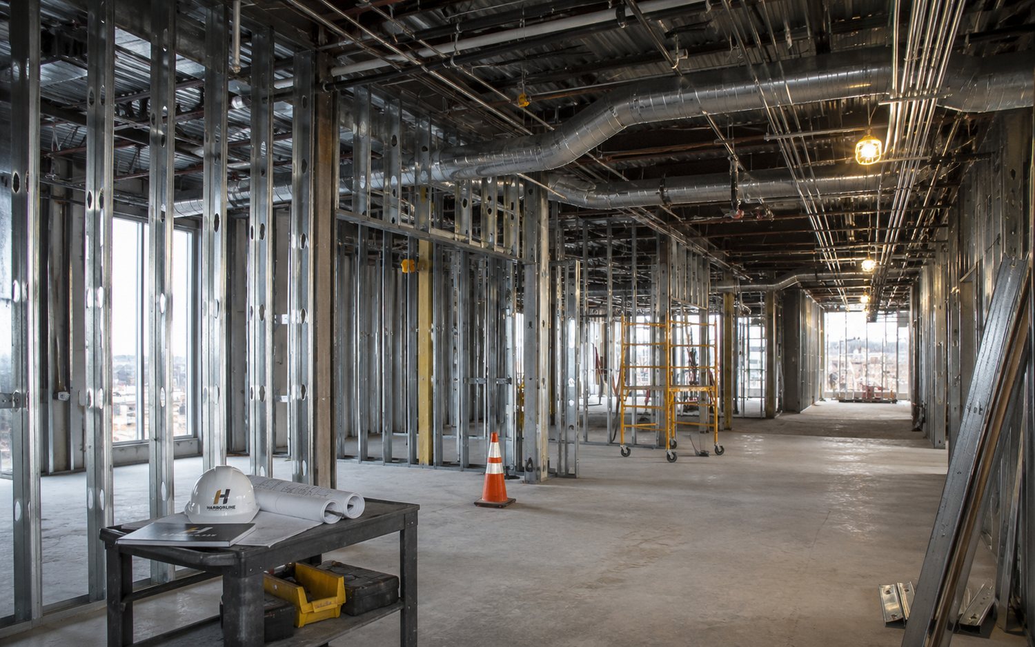 Commercial tenant space under construction with metal stud partitions, spiral ductwork, and overhead conduit, with drawings and a hard hat on a work cart